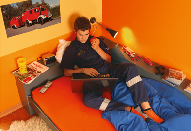 © Deutsche Jugendfeuerwehr Jugendlicher in Jugendfeuerwehrkleidung mit Laptop. Sein Zimmer ist ganz in Orangetönen gehalten.